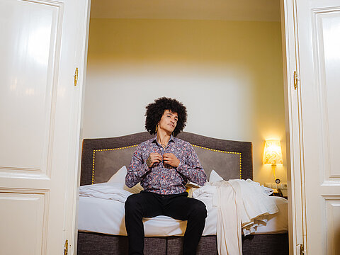 Mann im Blumenhemd knöpft sich auf dem Bett in stilvollem Hotelzimmer zu